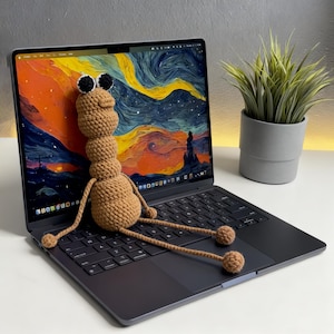 以下が含まれることがあります： 黒と白の目を持つ茶色の編み物のアリの人形が、ラップトップのキーボードの上に座っています。ラップトップの画面には、カラフルで渦巻く風景画が表示されています。ラップトップの右側には小さな鉢植えがあります。