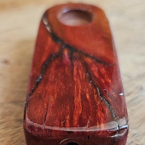 Puede incluir: Objeto rectangular de madera marrón rojizo con un agujero en un extremo. La madera tiene vetas oscuras y grietas. El objeto parece ser un artículo pequeño y portátil, posiblemente una pipa o un elemento decorativo.