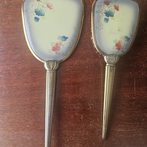May include: Two vintage vanity mirrors with floral designs. The mirrors have a gold-colored frame and handle. The mirror's surface is a pale yellow with a floral pattern in pink and blue. The handles are long and slender.
