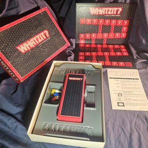 May include: A board game called "Whatzit?" is displayed, with its box and components visible. The box is red and black, with the game's title in white. The game board and pieces are red and black, and the instructions are also visible.