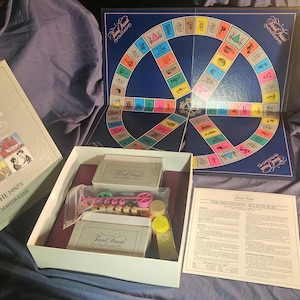 May include: An open box set of the Trivial Pursuit board game, featuring a blue game board with a colorful wheel design. The box and game components are visible, including cards and game pieces. The box reads "Trivial Pursuit" and "The 1980's Master Game."