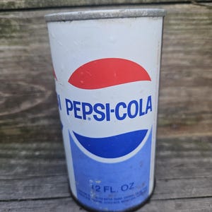 May include: Vintage Pepsi-Cola can with a white background, red and blue logo, and the words "PEPSI-COLA" in blue. The can has a 12 fl. oz. capacity and is slightly weathered, showing signs of age.