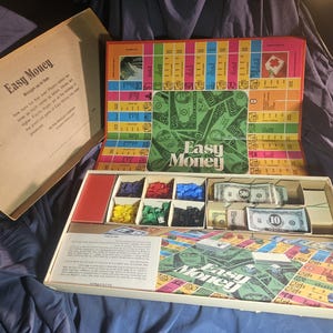 May include: Open vintage "Easy Money" board game. The game board is colorful with spaces for players to move around. The game includes play money, game pieces, and a box with the game's title. The box is open, revealing the game's contents.