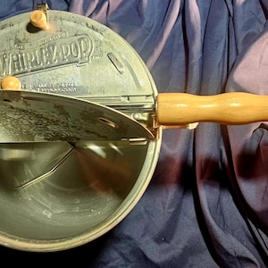 Peut inclure: Une machine à popcorn Whirley Pop vintage, avec un corps en métal argenté et une poignée en bois. Le couvercle est partiellement ouvert, révélant l'intérieur. La poignée et les boutons du couvercle sont en bois clair. Le texte "Whirley Pop" est visible.