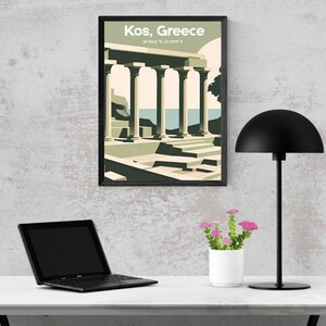 May include: A framed art print of Kos, Greece, featuring a minimalist illustration of ancient columns and architecture in shades of green, beige, and blue. The print is displayed above a white desk with a black laptop, a black desk lamp, and a small white pot of pink flowers.