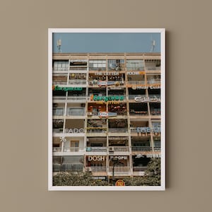 以下が含まれることがあります： The Letter、Kangaroo、1664、Pocha、Fado、Doshなどの様々な看板が掲げられた、複数のバルコニーを持つ多層階の建物ファサードの額入り縦型プリント。建物は格子状の建築デザインで構成されています。