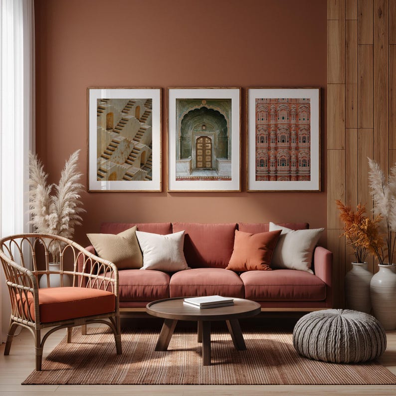 Op de afbeelding: Een woonkamerset bestaande uit een bank met drie kussens in een terracotta tint, een rotan fauteuil met bijpassend zitkussen, een ronde houten salontafel en drie ingelijste kunstwerken aan de muur met architecturale structuren.