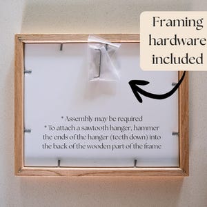 Op de afbeelding: Een rechthoekige fotolijst van lichtgekleurd hout, getoond vanaf de achterkant met metalen clips die de achterplaat vastzetten. Een klein plastic zakje met een zaagtandhanger is bevestigd. Tekst: Framing hardware included, Assembly may be required.