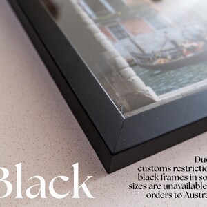 Op de afbeelding: Een zwarte fotolijst met verstekhoeken, met daarin een foto van een gondel op een kanaal. De tekst "Black" en "Due to customs restrictions, black frames in some sizes are unavailable for orders to Australia" is zichtbaar op de afbeelding.