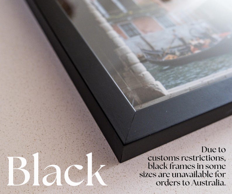 Op de afbeelding: Een zwarte fotolijst met verstekhoeken, met daarin een foto van een gondel op een kanaal. De tekst "Black" en "Due to customs restrictions, black frames in some sizes are unavailable for orders to Australia" is zichtbaar op de afbeelding.