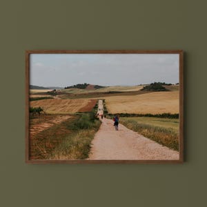 May include: A framed landscape photograph depicting a long, narrow dirt path winding through rolling golden wheat fields under a pale sky. A person with a blue backpack walks along the path, while other distant figures are visible further ahead.