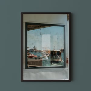 May include: Framed photograph of a Venetian canal scene. The image, viewed through a window, captures boats on the water, buildings, and a clear blue sky. The photograph is framed in a dark brown border.