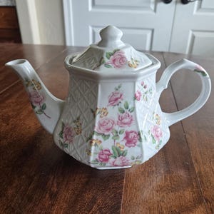 May include: White ceramic teapot with a floral rose pattern in pink and yellow. The teapot has a geometric design, a lid, and a curved handle. The teapot is sitting on a wooden table.