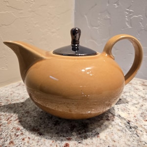 May include: A tan ceramic teapot with a black lid and handle. The teapot has a rounded body, a curved spout, and a loop handle. The lid has a small, dark knob. The teapot is sitting on a speckled countertop.