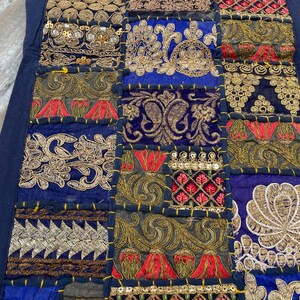 May include: A patchwork textile with a navy blue border. The design features squares of various fabrics in shades of blue, gold, red, and green, with intricate embroidered and sequined patterns. The overall aesthetic is ornate and decorative.
