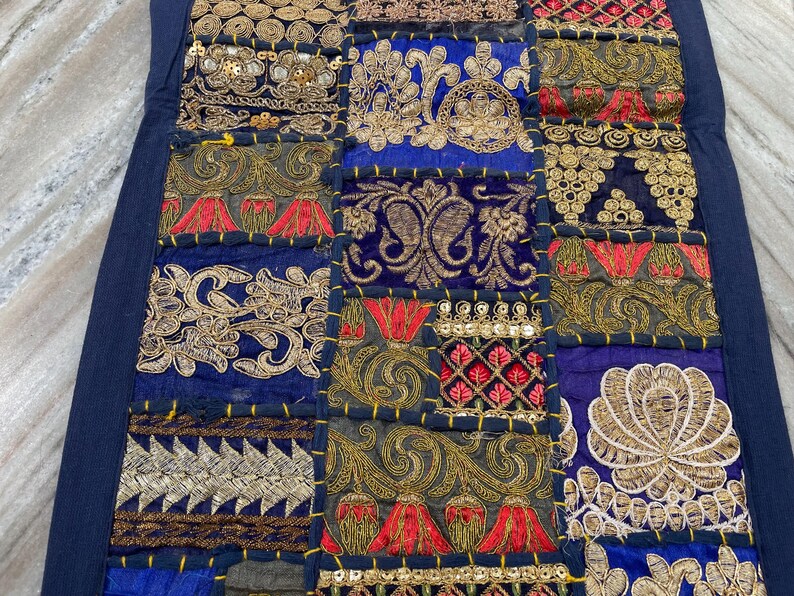 May include: A patchwork textile with a navy blue border. The design features squares of various fabrics in shades of blue, gold, red, and green, with intricate embroidered and sequined patterns. The overall aesthetic is ornate and decorative.