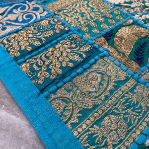 May include: Close-up of a patchwork textile with a teal and gold color scheme. The design features embroidered floral and geometric patterns. A teal border frames the top and left side of the textile.