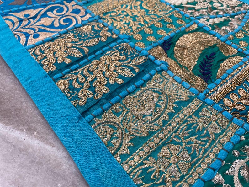 May include: Close-up of a patchwork textile with a teal and gold color scheme. The design features embroidered floral and geometric patterns. A teal border frames the top and left side of the textile.