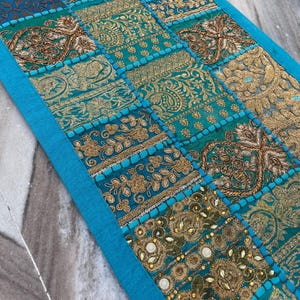 May include: A rectangular, turquoise and gold patchwork textile with intricate embroidered patterns. The design features various geometric shapes and floral motifs, with a bright blue border. The textile is likely used as a decorative table runner or wall hanging.