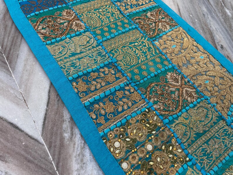 May include: A rectangular, turquoise and gold patchwork textile with intricate embroidered patterns. The design features various geometric shapes and floral motifs, with a bright blue border. The textile is likely used as a decorative table runner or wall hanging.