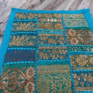 May include: A patchwork textile with a teal border and a variety of embroidered patterns. The central panel features squares of teal, gold, and blue fabrics with floral and geometric designs. The embroidery includes gold thread and beadwork.