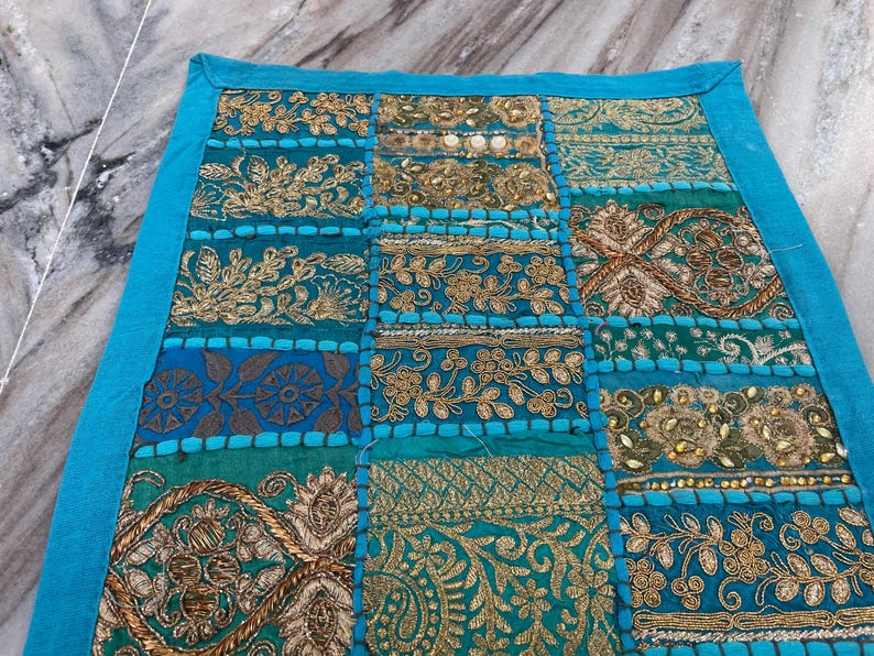 May include: A patchwork textile with a teal border and a variety of embroidered patterns. The central panel features squares of teal, gold, and blue fabrics with floral and geometric designs. The embroidery includes gold thread and beadwork.