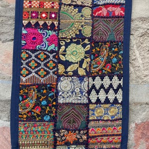 May include: A rectangular textile with a patchwork design. The patches feature intricate embroidery in various colors, including red, pink, gold, and blue, on a dark blue background. The edges are trimmed with a solid dark blue fabric.