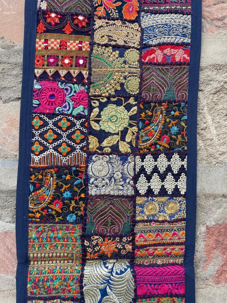 May include: A rectangular textile with a patchwork design. The patches feature intricate embroidery in various colors, including red, pink, gold, and blue, on a dark blue background. The edges are trimmed with a solid dark blue fabric.