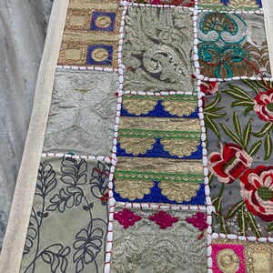 May include: A rectangular patchwork textile with a cream-colored border. The design features squares of various fabrics in shades of gold, silver, blue, green, and pink. Some squares have floral patterns, while others have embroidered details.