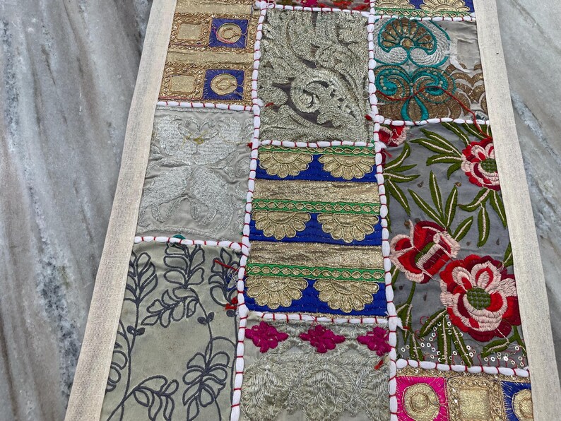 May include: A rectangular patchwork textile with a cream-colored border. The design features squares of various fabrics in shades of gold, silver, blue, green, and pink. Some squares have floral patterns, while others have embroidered details.