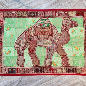 May include: A decorative textile featuring a camel design. The camel is composed of various fabric patches in shades of red, brown, and beige, set against a light green floral background. The border is a deep red, creating a striking contrast.