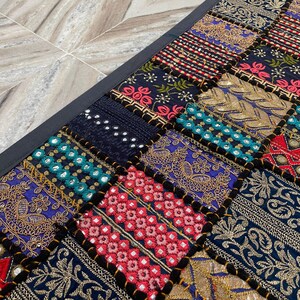 May include: A rectangular patchwork textile with a black border. The design features a variety of embroidered squares in shades of blue, red, gold, and teal. The squares have intricate patterns and textures.