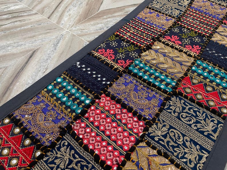 May include: A rectangular patchwork textile with a black border. The design features a variety of embroidered squares in shades of blue, red, gold, and teal. The squares have intricate patterns and textures.