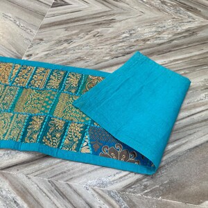 May include: A rectangular turquoise table runner with gold and teal patchwork designs. The runner is folded over, revealing a solid turquoise fabric on the right side. The patchwork features intricate patterns.