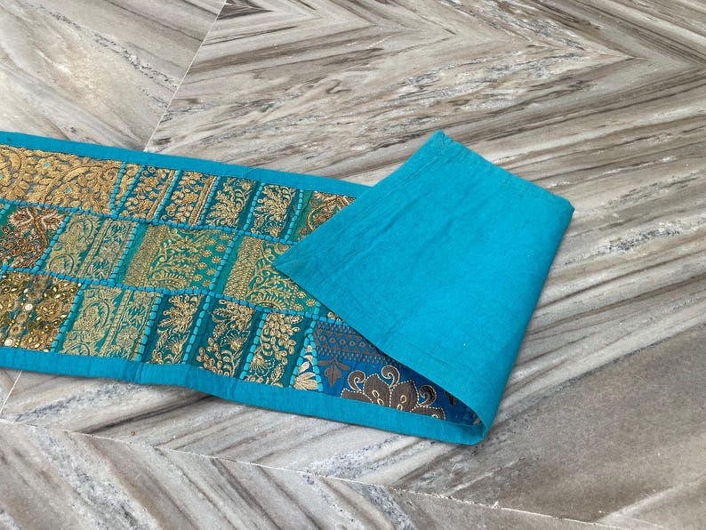May include: A rectangular turquoise table runner with gold and teal patchwork designs. The runner is folded over, revealing a solid turquoise fabric on the right side. The patchwork features intricate patterns.