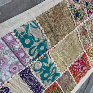 May include: A rectangular table runner with a patchwork design. The runner features various embroidered fabric squares in shades of gold, teal, pink, and purple. The edges are trimmed with a white border and red stitching.