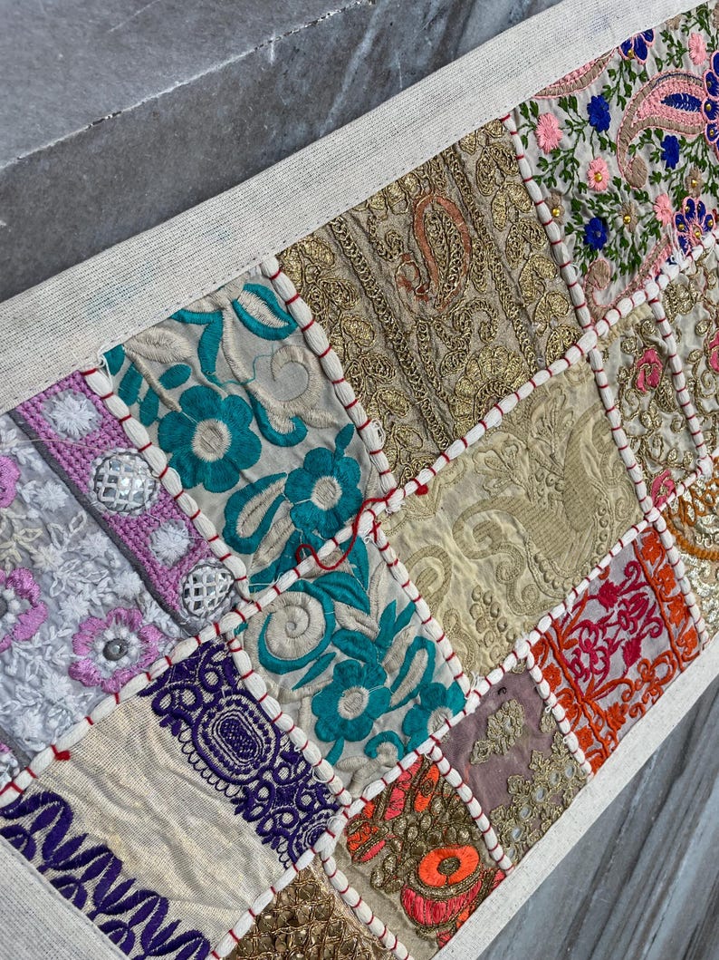 May include: A rectangular table runner with a patchwork design. The runner features various embroidered fabric squares in shades of gold, teal, pink, and purple. The edges are trimmed with a white border and red stitching.
