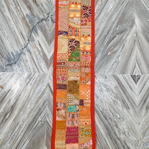 May include: A rectangular, patchwork textile with a vibrant mix of colors including orange, red, and gold. The design features a variety of patterns and textures, framed by a solid red border. Tassels hang from the bottom corners. This decorative piece is likely a table runner or wall hanging.