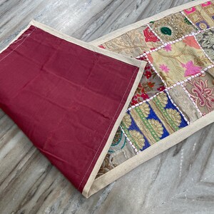 May include: A colorful patchwork table runner with a burgundy backing. The runner features a variety of patterned fabric squares in shades of pink, blue, green, and gold, bordered by a cream-colored fabric.