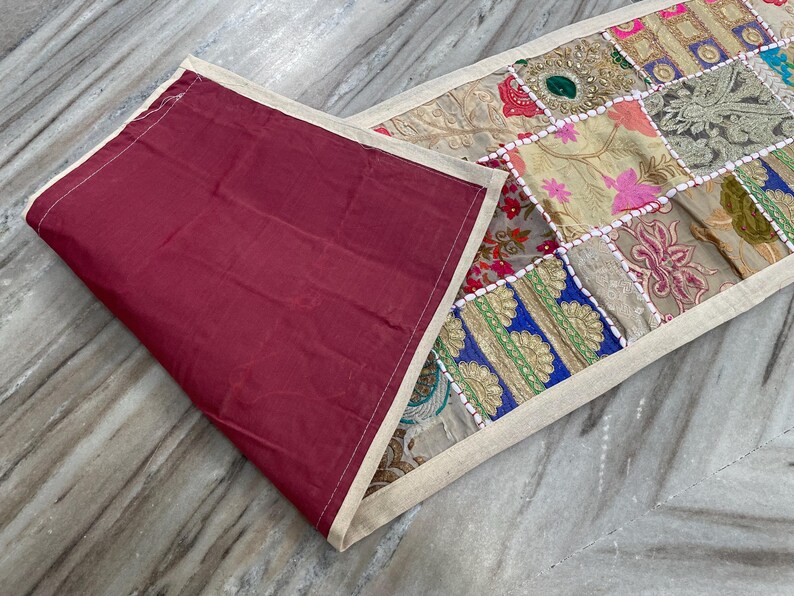 May include: A colorful patchwork table runner with a burgundy backing. The runner features a variety of patterned fabric squares in shades of pink, blue, green, and gold, bordered by a cream-colored fabric.