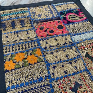 May include: A patchwork textile with a black border. The design features various embroidered squares in shades of blue, gold, orange, red, and pink. The embroidery includes floral and paisley patterns, with metallic gold thread and sequins.