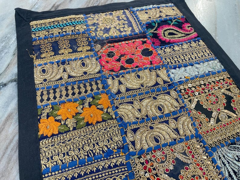 May include: A patchwork textile with a black border. The design features various embroidered squares in shades of blue, gold, orange, red, and pink. The embroidery includes floral and paisley patterns, with metallic gold thread and sequins.