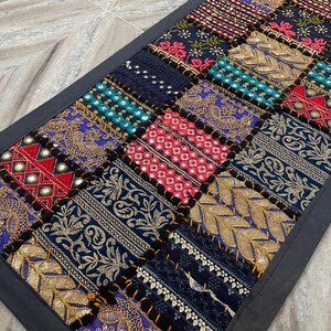 May include: A rectangular patchwork textile with a dark gray border. The patchwork design features squares of various colors and patterns, including gold embroidery, red and blue accents, and mirrored details. The textile is likely a decorative item.