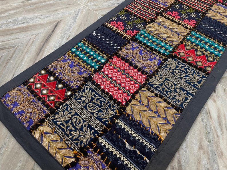 May include: A rectangular patchwork textile with a dark gray border. The patchwork design features squares of various colors and patterns, including gold embroidery, red and blue accents, and mirrored details. The textile is likely a decorative item.