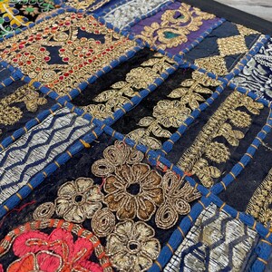 May include: Close-up of a patchwork textile with intricate embroidery. The design features a variety of geometric shapes and floral patterns in gold, red, and silver thread on a dark blue background. The patches are outlined with blue stitching.