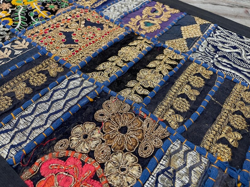 May include: Close-up of a patchwork textile with intricate embroidery. The design features a variety of geometric shapes and floral patterns in gold, red, and silver thread on a dark blue background. The patches are outlined with blue stitching.