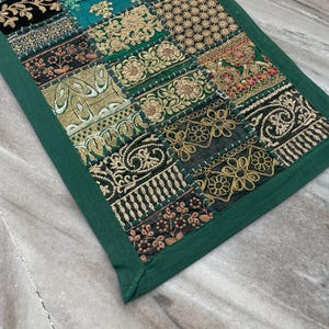May include: A rectangular patchwork textile with a green border. The patchwork features various embroidered patterns in gold, teal, black, and red. The textile is likely a decorative item, such as a table runner or wall hanging.