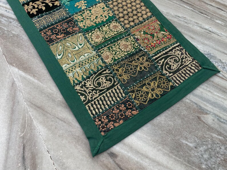 May include: A rectangular patchwork textile with a green border. The patchwork features various embroidered patterns in gold, teal, black, and red. The textile is likely a decorative item, such as a table runner or wall hanging.