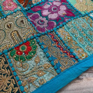 May include: Close-up of a patchwork textile with intricate embroidery. The design features squares of various colors, including teal, gold, red, and purple, with floral and paisley patterns. The edges are trimmed with a teal border.