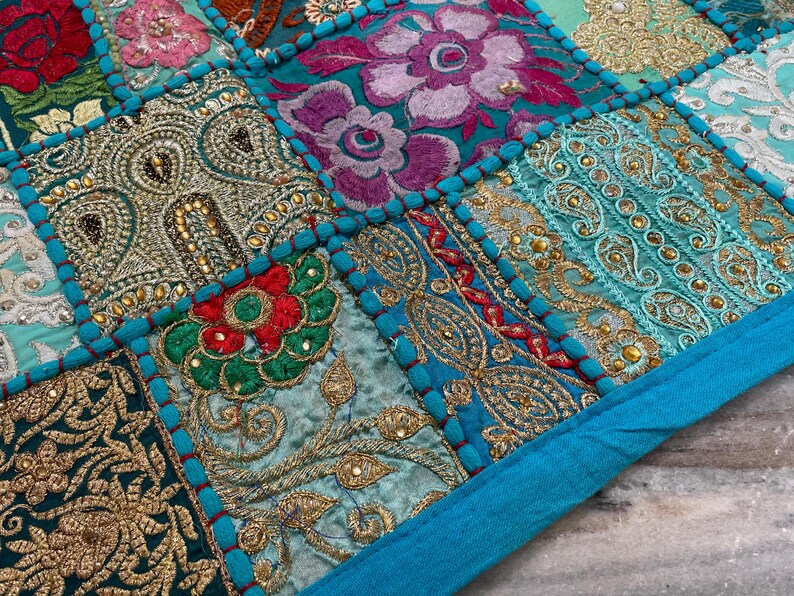 May include: Close-up of a patchwork textile with intricate embroidery. The design features squares of various colors, including teal, gold, red, and purple, with floral and paisley patterns. The edges are trimmed with a teal border.